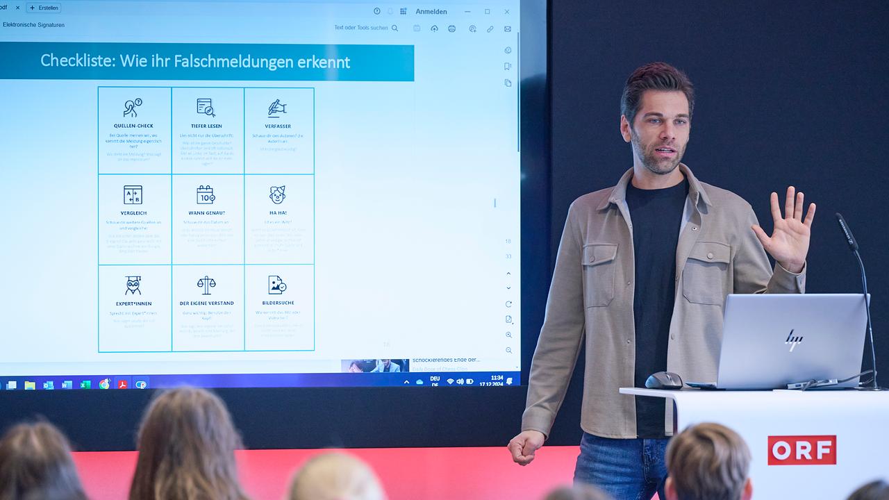 Ein Mann steht am Rednerpult mit seinem Laptop aufgeklappt und spricht zu den Schülern, von denen man nur die Hinterköpfe sieht. Hinter dem Redner ist eine Präsentation die zeigt: "Checkliste: Wie Ihr Falschmeldungen erkennt".