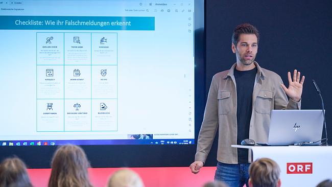 Ein Mann steht am Rednerpult mit seinem Laptop aufgeklappt und spricht zu den Schülern, von denen man nur die Hinterköpfe sieht. Hinter dem Redner ist eine Präsentation die zeigt: "Checkliste: Wie Ihr Falschmeldungen erkennt".